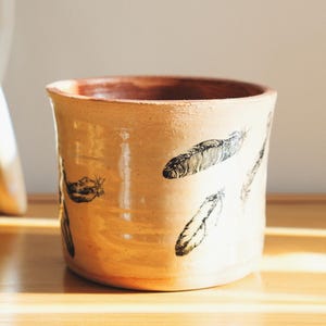 May include: A beige ceramic pot with a brown interior and black feather designs. The pot is cylindrical and sits on a wooden surface. A candle and decorative sculpture are in the background.