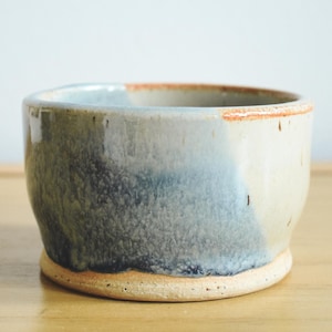 May include: A handmade ceramic bowl with a unique glaze. The bowl features a blend of colors, including blue, gray, and cream, with a textured appearance. The rim has a reddish-brown accent. The bowl sits on a light-colored base.