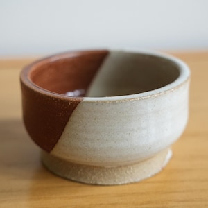 May include: A small, handcrafted ceramic bowl with a two-tone design. The upper portion is divided diagonally, featuring a warm brown and a creamy white. The bowl sits on a small, circular base, showcasing a minimalist aesthetic.