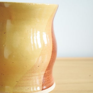 May include: A ceramic mug with a yellow, orange, and brown glaze. The mug has a rounded shape and a slightly flared lip.