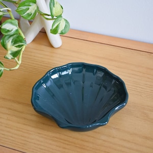 May include: A dark teal ceramic dish shaped like a seashell. The dish is sitting on a wooden surface.