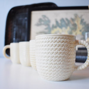 May include: Four white ceramic mugs with a textured, geometric pattern. The mugs have a rounded shape and a handle on the side. The mugs are arranged in a row on a white surface.