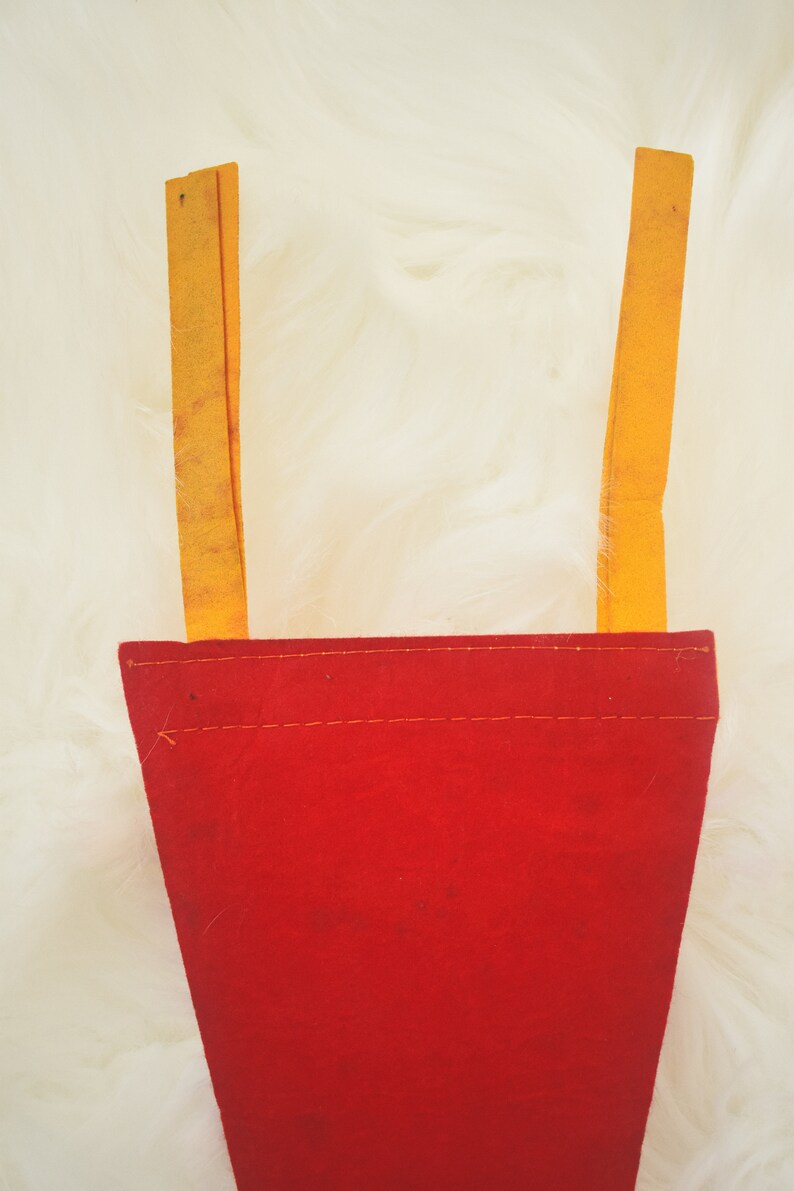 May include: A red felt pennant with two yellow straps. The pennant has a rectangular shape with a stitched seam along the top. The yellow straps are attached to the top of the pennant.