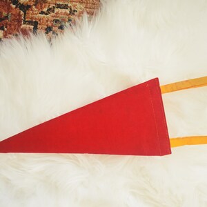 May include: A red felt pennant with a triangular shape. The pennant has two yellow wooden dowels attached to the back. The pennant is displayed on a white, fluffy surface.