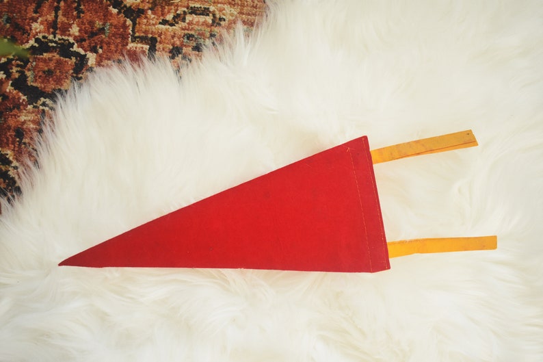 May include: A red felt pennant with a triangular shape. The pennant has two yellow wooden dowels attached to the back. The pennant is displayed on a white, fluffy surface.