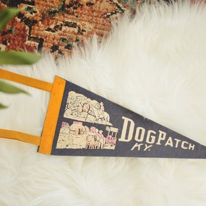 May include: A navy blue pennant with the text "Dogpatch KY" in a serif font. The pennant has a yellow border and two yellow tabs. The pennant features a graphic of a town.
