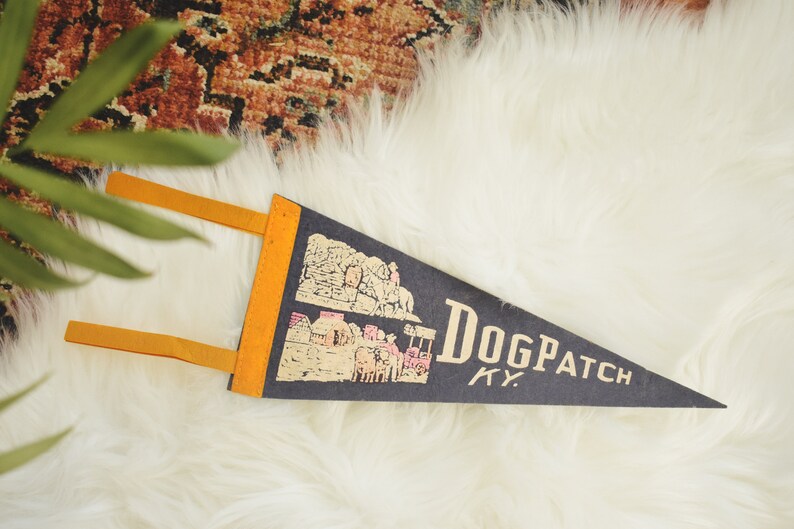 May include: A navy blue pennant with the text "Dogpatch KY" in a serif font. The pennant has a yellow border and two yellow tabs. The pennant features a graphic of a town.