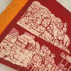 May include: A vintage red felt pennant with an orange border. The pennant features cream-colored illustrations of a mountain scene, a building, and figures. Text includes "MOONSHINE STAGE" and "DOGPATCH".