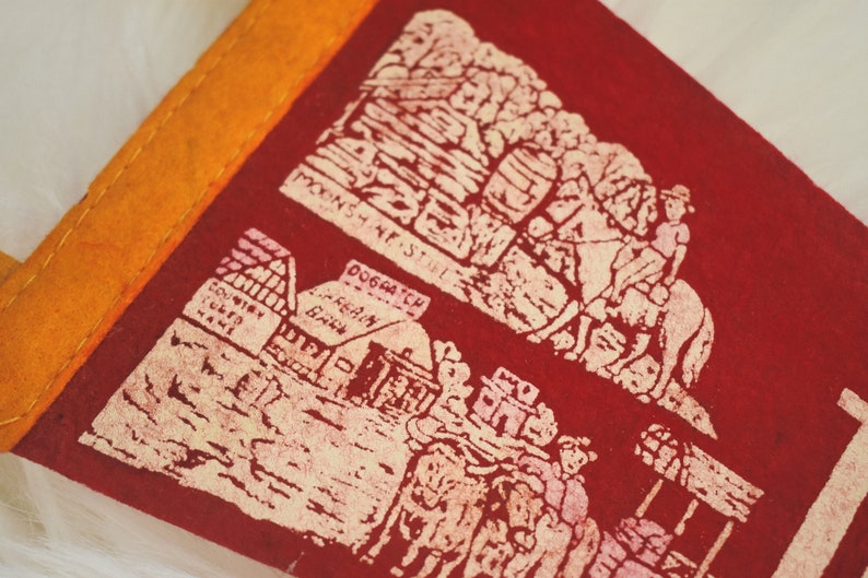 May include: A vintage red felt pennant with an orange border. The pennant features cream-colored illustrations of a mountain scene, a building, and figures. Text includes "MOONSHINE STAGE" and "DOGPATCH".