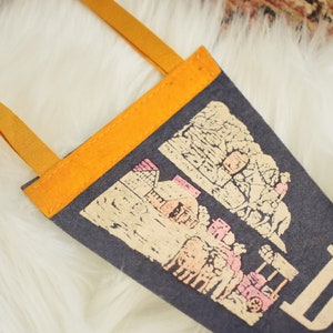 May include: A dark gray felt pennant with a tan and pink illustration of a town and the word "Do" in tan. The pennant has a bright orange border and tan straps for hanging. The pennant is displayed on a white, fluffy surface.