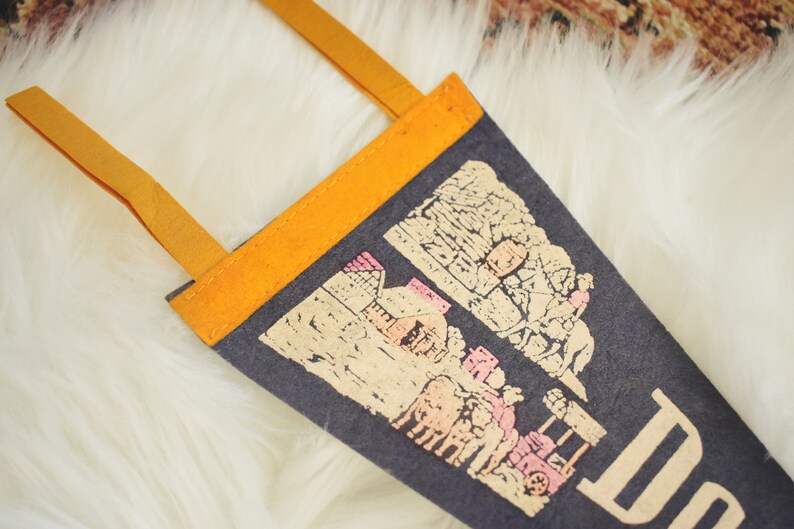 May include: A dark gray felt pennant with a tan and pink illustration of a town and the word "Do" in tan. The pennant has a bright orange border and tan straps for hanging. The pennant is displayed on a white, fluffy surface.