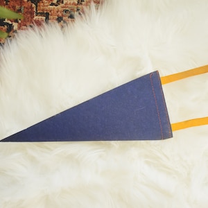 May include: A navy blue pennant with orange accents. The pennant is triangular and has orange tabs extending from the top. The pennant is lying on a white, fluffy surface.