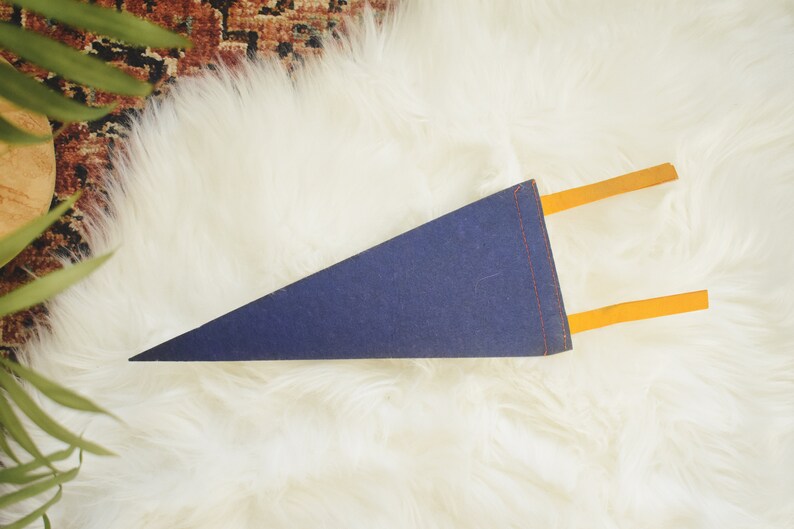 May include: A navy blue pennant with orange accents. The pennant is triangular and has orange tabs extending from the top. The pennant is lying on a white, fluffy surface.