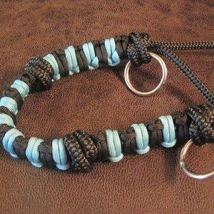 Könnte beinhalten: Ein schwarzes und hellblaues Paracord-Halsband für Hunde mit zwei silbernen Metallringen. Das Halsband besteht aus gewebtem schwarzen und hellblauen Kordel, mit einer Reihe von Knoten und Schlaufen. Der Hintergrund ist eine braune lederartige Oberfläche.