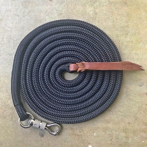 Op de afbeelding: Zwarte touwhalster met een bruin leren handvat en een zilveren karabijnhaak. Het touw is in een cirkel gewikkeld.