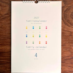 Könnte beinhalten: Ein Wandkalender für 2027 mit weißem Deckblatt und einer Drahtbindung am oberen Rand. Der Text lautet 2027 familienkalender mit ferienterminen, family calendar with german school holidays und 4 persons. Er zeigt ein Raster aus kleinen, bunten Punkten.