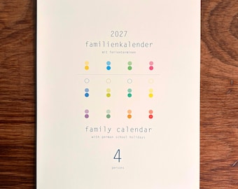 May include: A 2027 wall calendar with a white cover, wire-bound at the top. The text reads 2027 familienkalender mit ferienterminen, family calendar with german school holidays, and 4 persons. It features a grid of small, colorful dots.