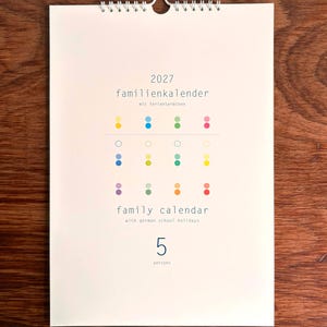 Könnte beinhalten: Ein Wandkalender für 2027 mit weißen Seiten und einer silbernen Drahtspirale. Das Deckblatt zeigt den Text 2027 familienkalender mit ferienterminen, family calendar with german school holidays und 5 persons sowie ein Raster aus bunten Punkten.
