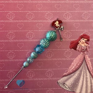 以下が含まれることがあります： 銀色のフォーク、緑と白のパターンビーズ、赤と白のアリエルチャームが付いた、ターコイズブルーとブルーのビーズチャーム。チャームは、白いハート柄のピンクの背景の上にあります。