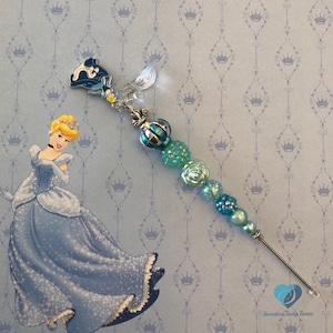 May include: A decorative pen with a Cinderella theme. The pen features a blue and silver design with various beads and charms. The background is light blue with a Cinderella illustration and crown pattern.
