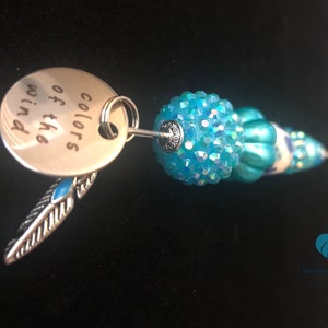 以下が含まれることがあります： 「風の色の」と書かれた銀色のチャームが付いたターコイズブルーのビーズキーホルダー。チャームは銀色の羽根チャームに繋がっています。