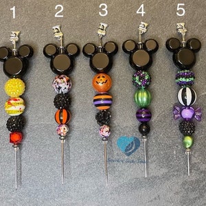 以下が含まれることがあります： 黒いミッキーマウスの頭の飾りが付いた、5つの装飾的なカクテルピック。各ピックは、カボチャ、ストライプ、キャンディーなど、さまざまなデザインのカラフルなビーズが特徴です。ハロウィーンやテーマパーティーに最適です。