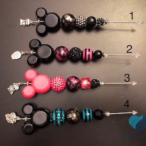 以下が含まれることがあります： さまざまなデザインのビーズペン先が4つ。1つ目は黒いビーズ、2つ目はピンクと黒のビーズ、3つ目はピンクのビーズ、4つ目は青と黒のビーズです。各ペン先には、銀色の金属製の留め金とペンの先端が付いています。