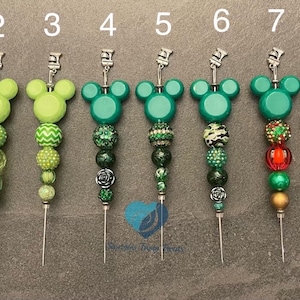 以下が含まれることがあります： ミッキーマウスの形をしたトップが付いた、8本の装飾的な縫い針のコレクション。針は、さまざまな緑色とカラフルなビーズのデザインが特徴です。各針は銀色のトップと鋭い尖った端を持っています。