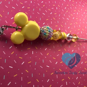 以下が含まれることがあります： 黄色のミッキーマウスの頭のチャームに銀色のチャームが取り付けられています。チャームは、黄色、白、青のビーズでできたビーズネックレスに糸で通されています。