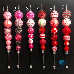 May include: Six Valentine's Day themed beaded keychains with different designs. Each keychain features a variety of beads in pink, red, and white with different patterns and textures. The keychains are numbered 1 through 6.