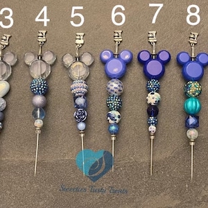 以下が含まれることがあります： 様々な青いビーズのデザインとミッキーマウスの頭の飾りが付いた、10個の装飾ピンのコレクション。ピンは透明、模様入り、愛国的なビーズが特徴です。ピンは灰色の表面に表示されています。各ピンの長さは約10cmです。