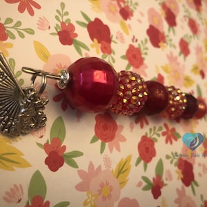 以下が含まれることがあります： 扇形のチャームが付いた赤と銀のビーズのヘアースティック。ビーズは丸型、ファセットカット、キラキラなど、さまざまなサイズと形をしています。扇形のチャームは銀色で、細かなデザインが施されています。