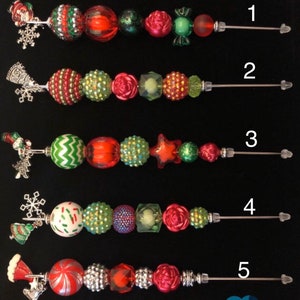 以下が含まれることがあります： さまざまな色のビーズとチャームが付いた、5つのクリスマスをテーマにした装飾串。各串には、赤、緑、白、銀色のビーズと、雪の結晶やサンタの帽子などのお祝いのチャームが組み合わされています。串は黒い背景に配置されています。
