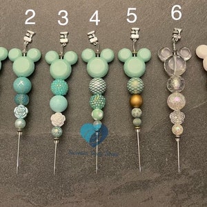 以下が含まれることがあります： ミッキーマウスの頭のデザインが施された、7つの装飾的な針止め。各針止めは、ミントグリーン、ティール、ゴールド、ホワイトの色合いのビーズの異なる組み合わせが特徴です。針止めはグレーの表面に表示されています。