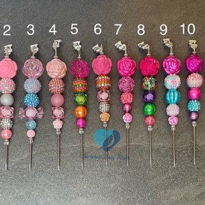 Puede incluir: Once bolígrafos decorativos con cuentas en varios tonos de rosa, con puntas de metal plateado. Cada bolígrafo presenta una combinación única de cuentas, incluyendo diseños redondos, facetados y florales. Los bolígrafos están dispuestos verticalmente sobre un fondo gris.