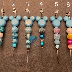 以下が含まれることがあります： 水色のミッキーマウスの頭のデザインが施された、6つの装飾的な帽子ピン。各ピンは、青、ピンク、オレンジ、白の色合いのカラフルなビーズの異なる組み合わせが特徴です。ピンは銀色の金属針が付いています。