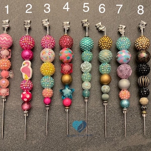 以下が含まれることがあります： 異なるデザインのカラフルなビーズキーホルダーが8つ。フラミンゴ、人魚、星などがあります。各キーホルダーは、シルバーのクリップと細い金属棒に取り付けられています。