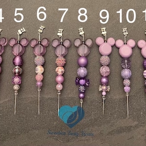 Peut inclure: 14 pinces à cheveux en perles violettes et roses avec un motif de tête de Mickey Mouse. Les pinces sont numérotées de 1 à 14.