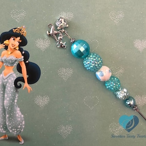 Pode incluir: Um bracelete de charme turquesa e prata com um charme de prata da lâmpada do gênio de Aladdin e uma conta turquesa. O bracelete está sobre um fundo verde com corações brancos.