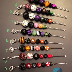 以下が含まれることがあります： 魔女の帽子、カボチャ、幽霊など、さまざまなチャームが付いた、ハロウィンをテーマにしたビーズキーホルダーが9個あります。ビーズは、黒、オレンジ、緑、紫など、さまざまな色です。