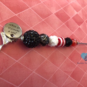 以下が含まれることがあります： スプーンと「Just 4 spoons of sugar」というテキストが刻まれたシルバーチャームが、黒、白、赤のビーズでできたネックレスに付いている。