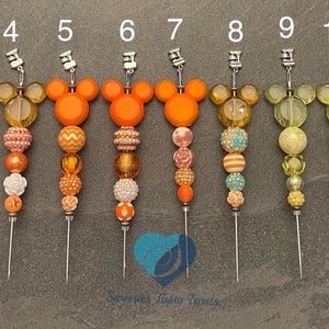 以下が含まれることがあります： ミッキーマウスの頭のデザインが施された、12個の装飾ケーキトッパーのコレクション。各トッパーは、銀色の金属針と、オレンジ、黄色、ピンクの色合いのカラフルなビーズの茎が特徴です。トッパーは灰色の表面に配置されています。