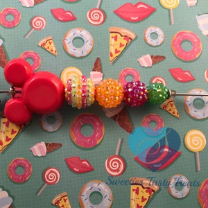 以下が含まれることがあります： 赤いデザインのビーズペン。黄色、オレンジ、ピンク、緑の色合いのカラフルでキラキラしたビーズが続きます。ミキサーの小さなシルバーチャームがペンからぶら下がっています。背景は、ドーナツ、ピザスライス、アイスクリームコーンのティールパターンです。