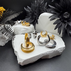 May include: A collection of C-shaped ear cuffs in polished gold and silver tones. These circular jewelry pieces feature a smooth, thick band design with an open gap, displayed in various sizes across multiple white stone-like platforms.