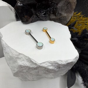 May include: Two straight barbell body jewelry pieces featuring round, iridescent white opal-like cabochon ends. One barbell has a silver-toned metal finish, while the other has a gold-toned metal finish. Both items feature two circular settings.