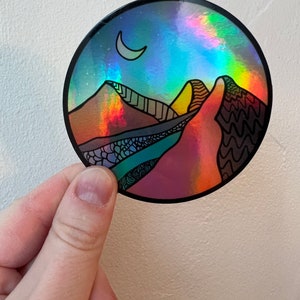 Può includere: Un adesivo olografico rotondo raffigurante un paesaggio desertico con montagne, una luna crescente e un cielo stellato. L'adesivo ha un bordo nero ed è tenuto da una mano su uno sfondo bianco. Il design incorpora vari colori.