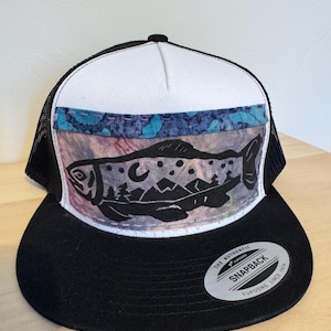 May include: A black and white trucker hat with a fish graphic. The hat features a black brim, a white front panel, and a black mesh back. The fish design is black and includes a mountain and moon scene. The hat has a Snapback closure.