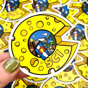 May include: A yellow sticker in the shape of a cheese wedge with the letters "SCI" and a Colorado flag. The center of the cheese wedge features a landscape scene with mountains and buildings. The sticker has a white border.