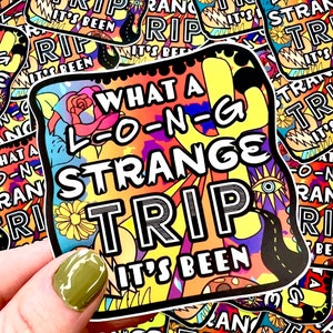 May include: A colorful sticker with the text "WHAT A L-O-N-G STRANGE TRIP IT'S BEEN" in a bold, outlined font. The background features a mix of floral and psychedelic designs in yellow, orange, purple, and blue. The sticker is held by a hand with a green painted nail.
