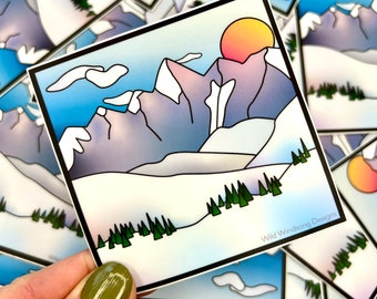 May include: Square sticker featuring a mountain landscape with a sunset. The design includes snow-capped mountains, green trees, and a gradient sky. The text "Wild Windsong Designs" is printed on the bottom right corner.
